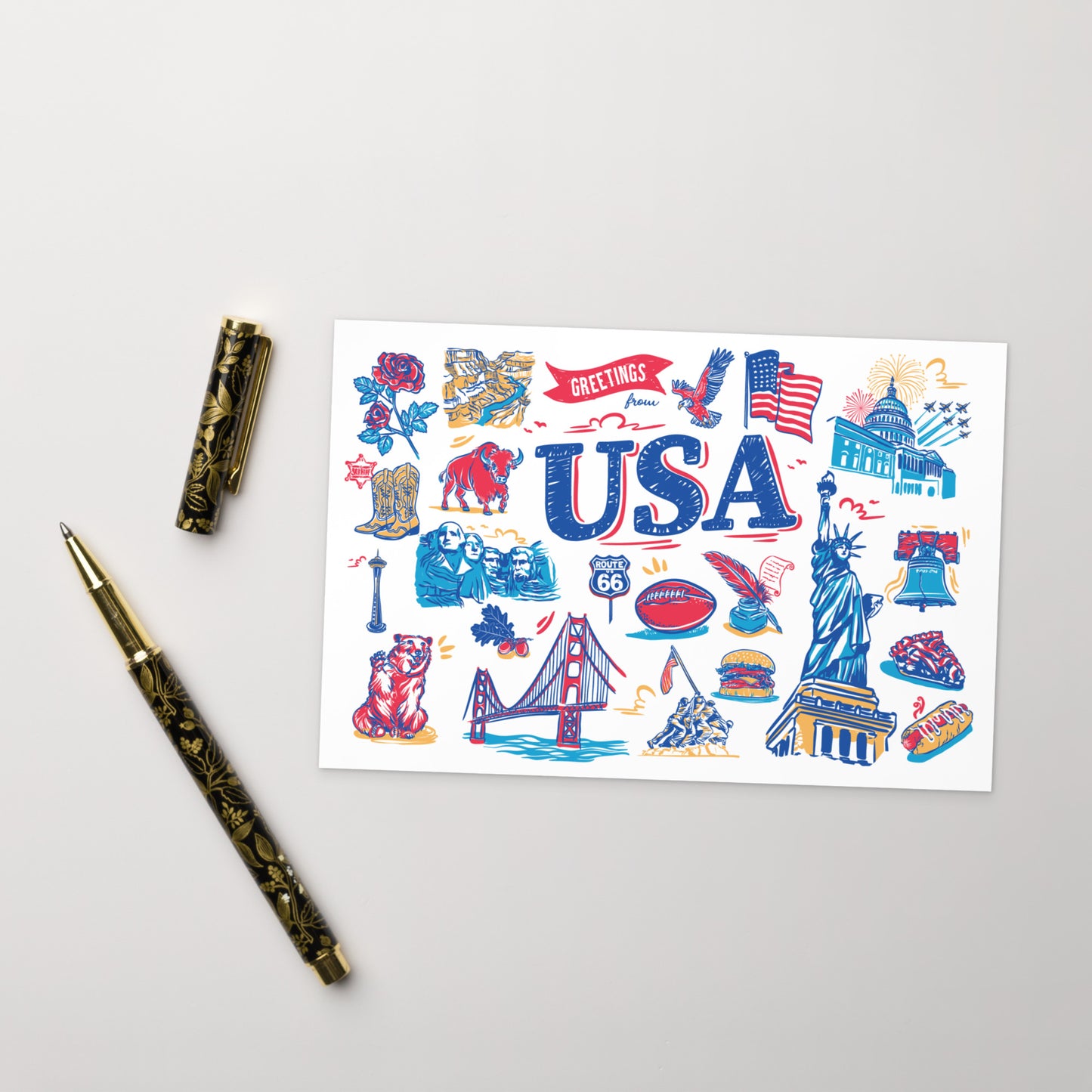 A white rectangular art print featuring colorful USA-themed illustrations and the word 'USA' in large blue letters, styled on a light gray surface with a gold-and-black pen and its cap beside it.