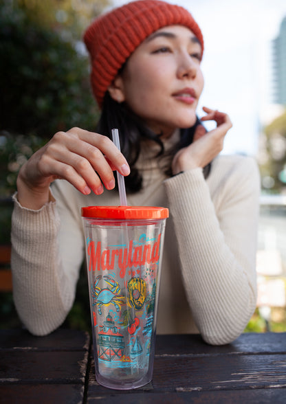 Person drinking from a colorful tumbler with 'Maryland' text and crab design, wearing a red beanie.