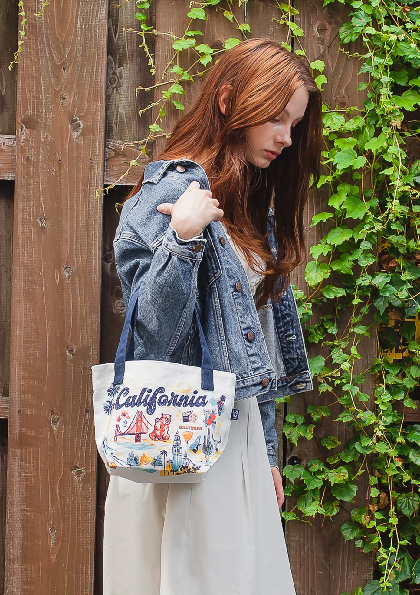 Woman holding a tote bag with 'California' text against a wooden wall with greenery