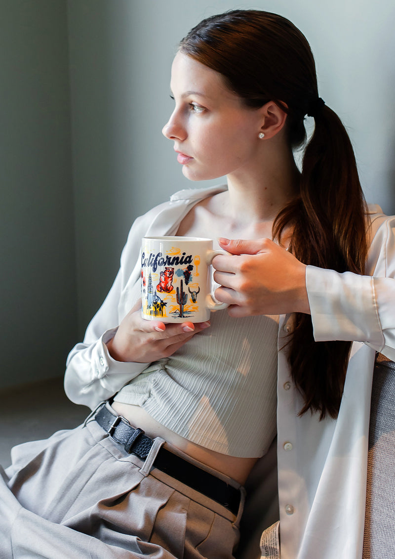 Woman holding a mug with a 'California' design, sitting indoors.