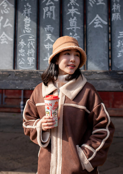 A woman wearing a brown shearling-lined jacket with beige trim and a matching beige bucket hat, holding a red cup in front of a dark wooden wall with Chinese calligraphy.