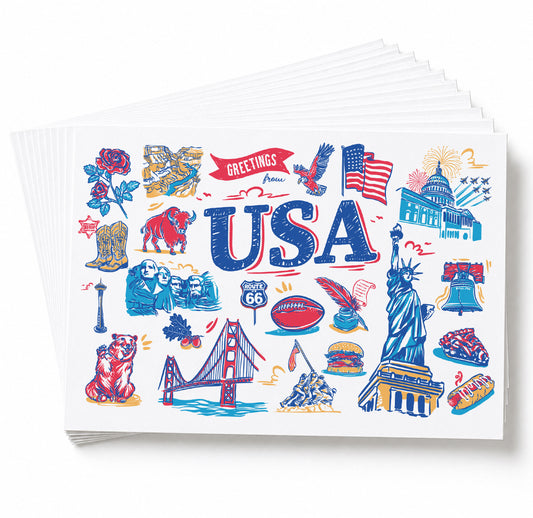 A stack of white greeting cards fanned out on a white background, with the top card featuring a colorful USA-themed illustration including the word 'USA,' the Statue of Liberty, the Golden Gate Bridge, the American flag, and other patriotic icons.