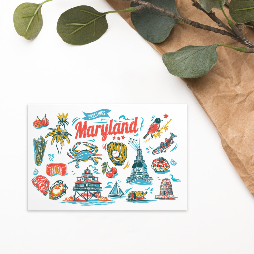 A white greeting card featuring a colorful hand-drawn illustration of Maryland landmarks, food, and nature, styled on a white surface with brown paper and green leaves.