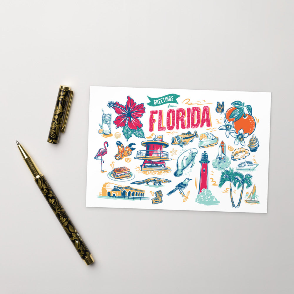 A flat-lay photograph of a 'Greetings Florida' art print card, styled with a gold-patterned pen and pen cap on a light gray background. The card features colorful hand-drawn illustrations of Florida landmarks, wildlife, and culture, including a lighthouse, flamingo, orange, and palm trees.