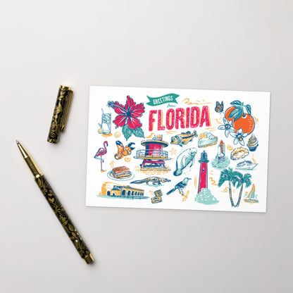 A flat-lay photograph of a 'Greetings Florida' art print card, styled with a gold-patterned pen and pen cap on a light gray background. The card features colorful hand-drawn illustrations of Florida landmarks, wildlife, and culture, including a lighthouse, flamingo, orange, and palm trees.