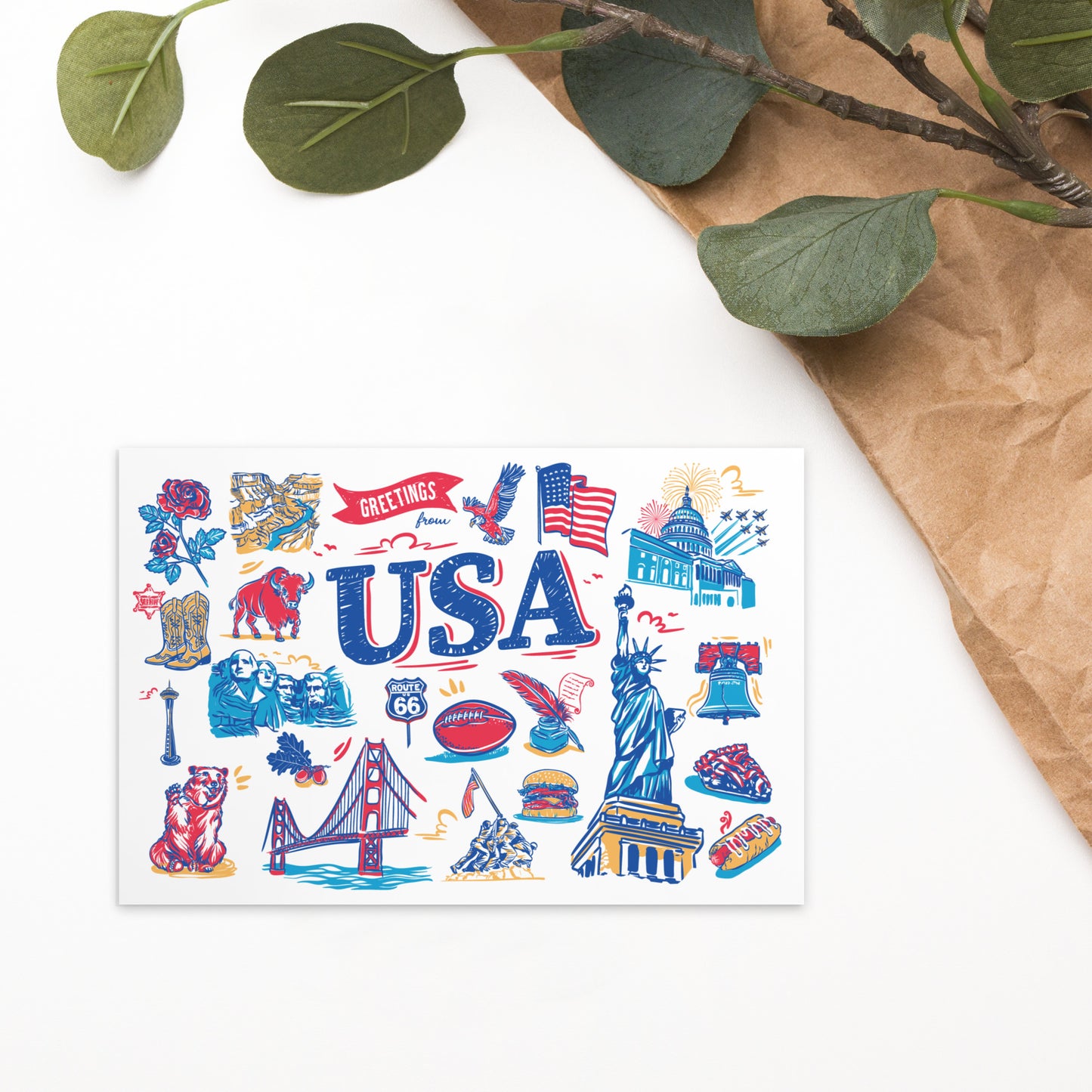 A USA-themed art print or postcard featuring colorful illustrations of American landmarks, symbols, and words like 'USA' and 'Greetings,' styled on a white surface with brown paper and green leaves in the corner.