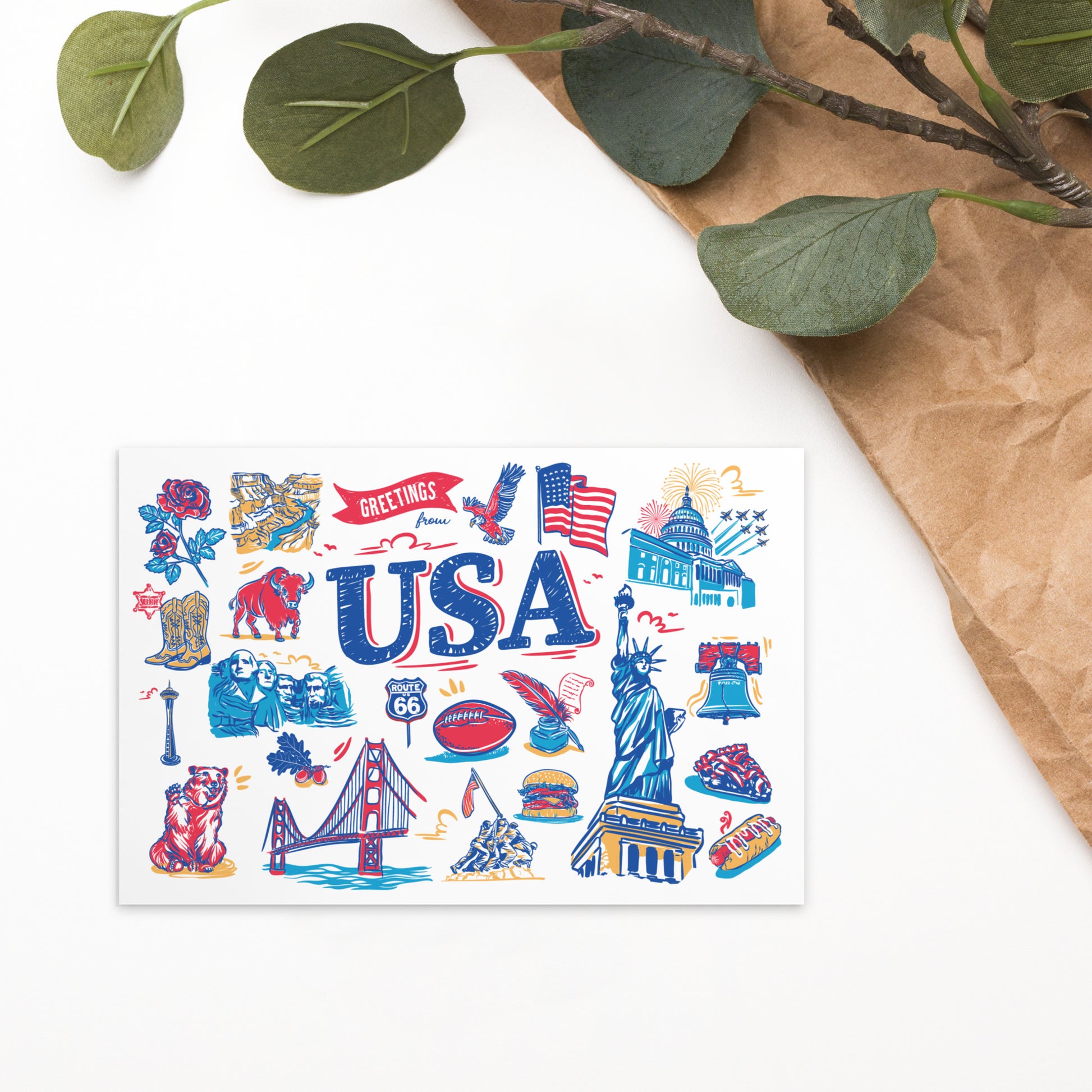 A USA-themed art print or postcard featuring colorful illustrations of American landmarks, symbols, and words like 'USA' and 'Greetings,' styled on a white surface with brown paper and green leaves in the corner.